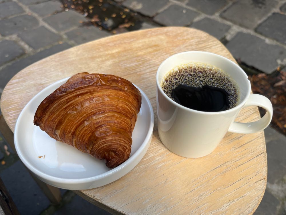 A plain croissant sitting on a white ceramic plate, next to a cup of filter coffee
