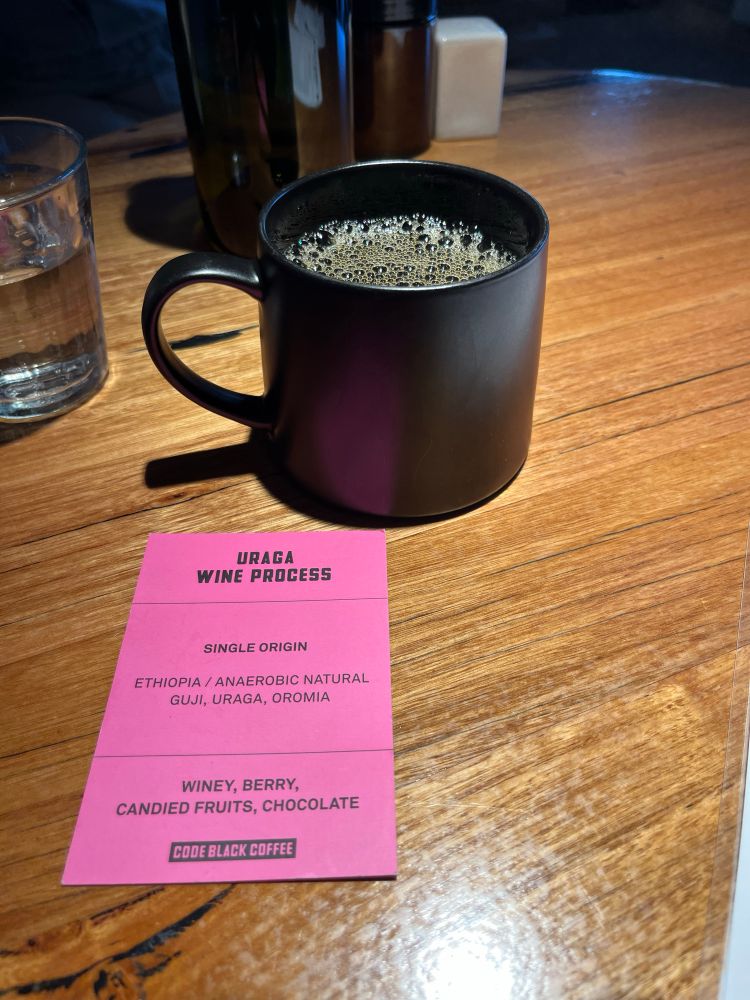 A cup of filter coffee with a card explaining the kind of beans used to make it. Card says “Uganda Wine Process — Single origin, Ethiopia / Anaerobic natural guji, uraga, oromia — Winey, berry candied fruits, chocolate