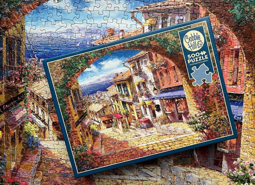 A Cobble Hill 500 piece puzzle box on top of a  completed puzzle of a view of Arch De Cagnes in France. It shows an archway covered in foliage that opens up to a view of a cobble stone street with boutique shops flanking it. The stairs lead down. And in the distance are buildings that look like homes with a view of the ocean and a snow capped mountain. 