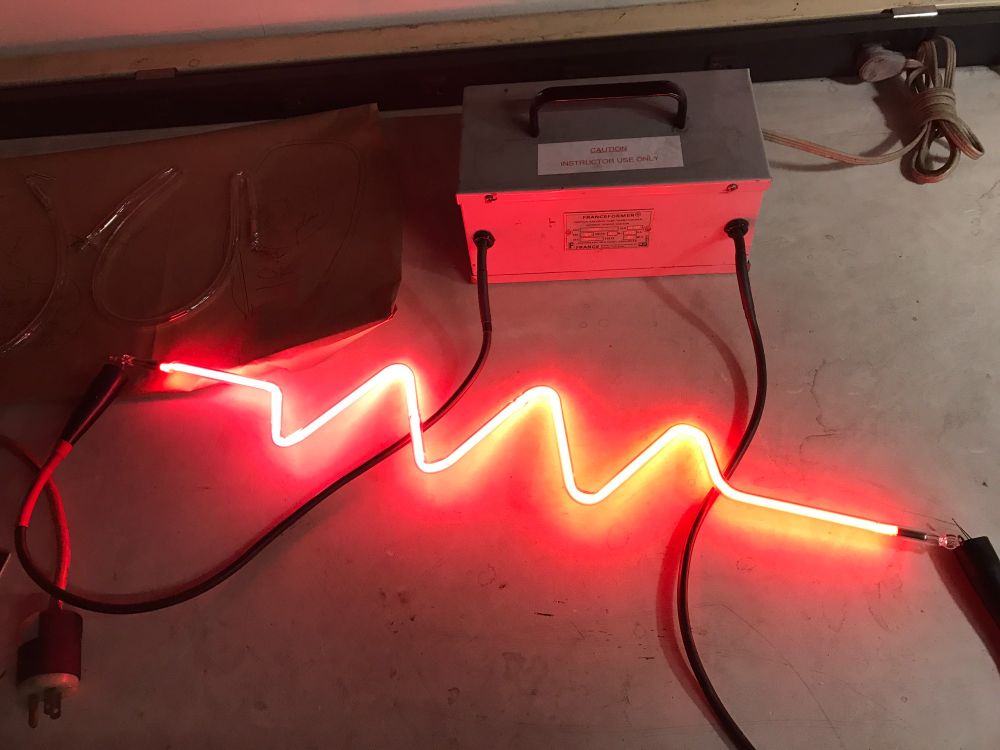 A piece of neon tubing shaped into the electronic symbol for a resistor, sitting on a workbench and hooked up to a high voltage power supply so that it glows bright red.