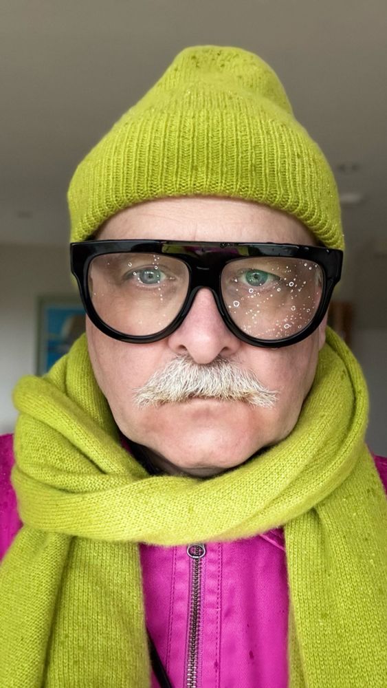 A man with a moustache in large black framed glasses wearing a likes green hat and scarf and a pink jacket 