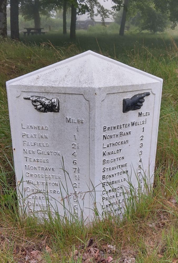 Cast iron milestone, painted white with black text wearing off and two pointing hands left and right. Destinations include: Lawhead, Peat Inn, Brewester Wells, New Gilston, Lathockar.