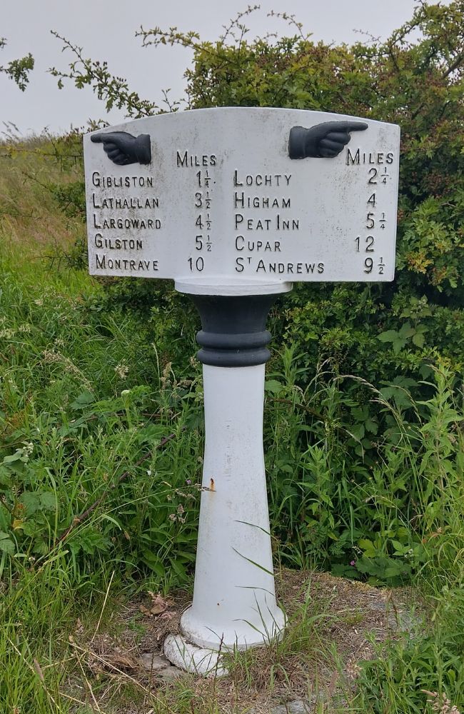 Unusual cast iron milestone on a pillar, painted black and white. Destinations include Gibliston, Lochty, Peat Inn, Cupar, St Andrews