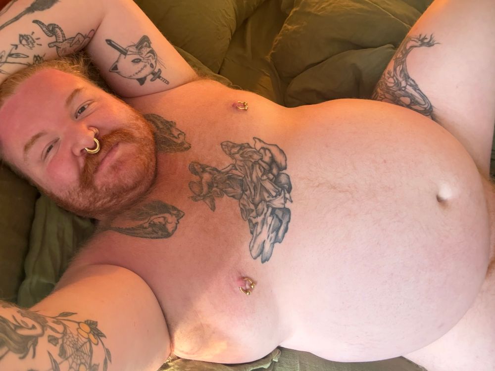 Selfie of Dave laying back on a green bed, naked and visible from just below their belly up. They have tattoos on their arms, legs, and chest, with pierced nipples and a septum ring. They have ginger hair, a scruffy beard, and light colored body hair.