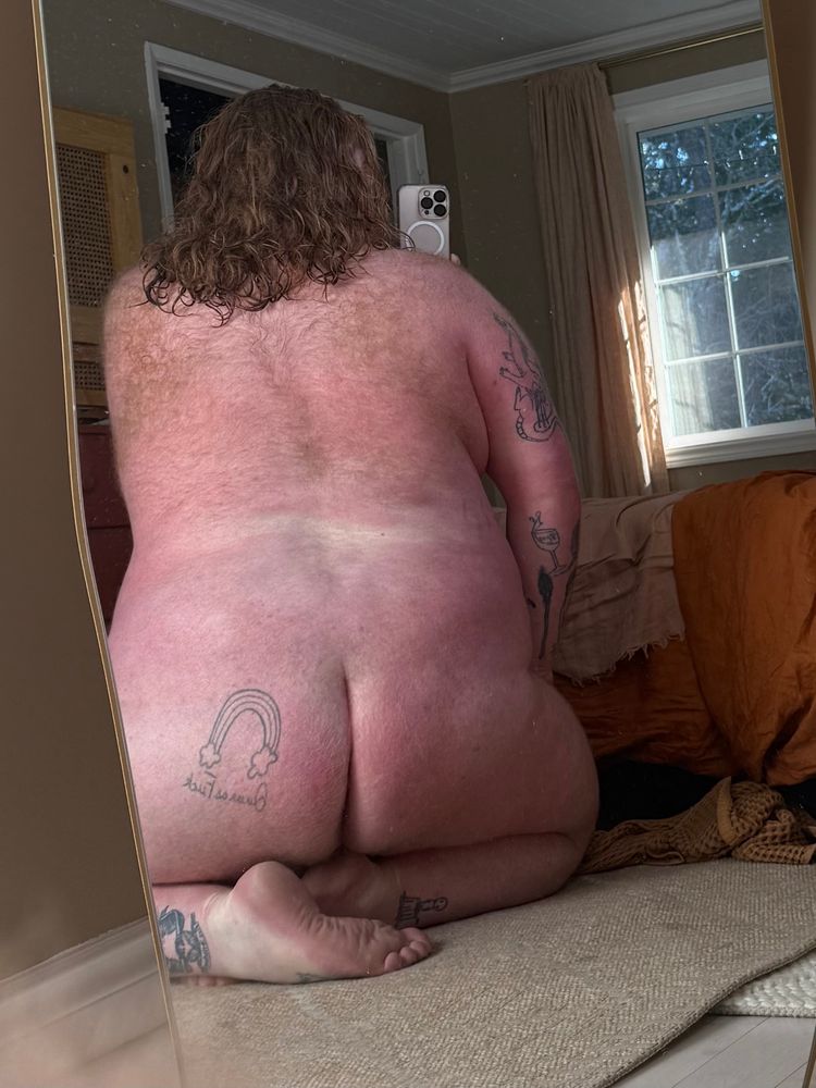 Picture of Dave from behind. They’re kneeling and you can see tattoos on their legs, ass, and arm. They’re hair is red, curly, and wet, and falls past their shoulders. They have hairy shoulders and back. 