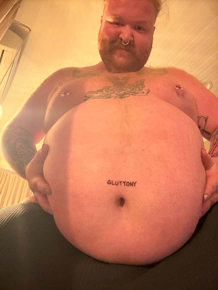 Picture of Dave holding their round gut. The word gluttony is tattooed above their belly button.