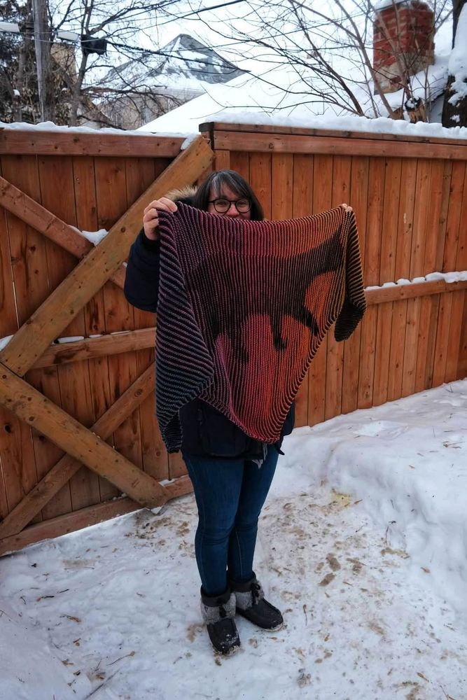 Woman in front of a fence holding up a blue, red, yellow shawl with a black cat on it. Ther is snow on the ground.