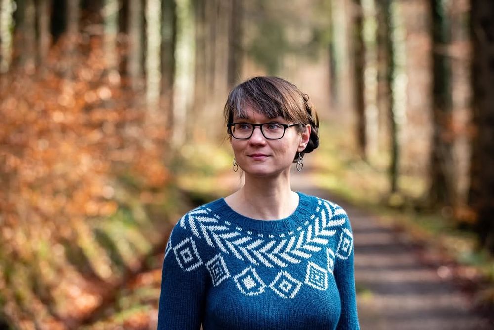 Blue sweater with white geometric pattern around the yoke worn by a brown-haired woman with black glasses.