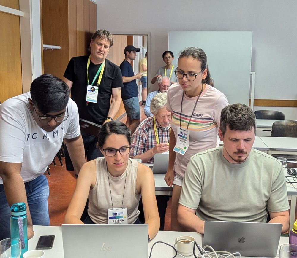 Lorena is sitting at a table in front her laptop looking intently at the screen. She has two collaborators peering over her shoulder and one sitting beside working in parallel.