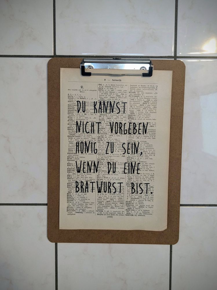 An einer gefliesten Wand hängt ein Klemmbrett. Darauf ist eine alte Lexikonseite befestigt, auf der handschriftlich drübergeschrieben steht: Du kannst nicht vorgeben, Honig zu sein, wenn du eine Bratwurst bist.