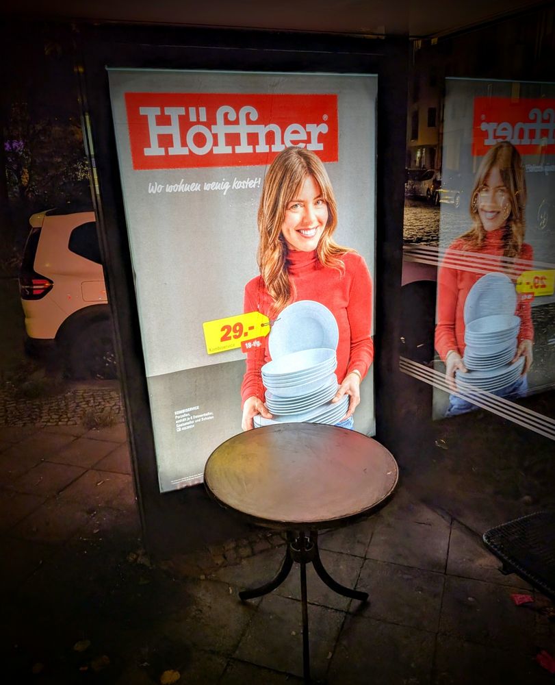 Vor einem erleuchteten Höffner-Plakat mit einer Frau die Teller trägt steht ein Holztisch