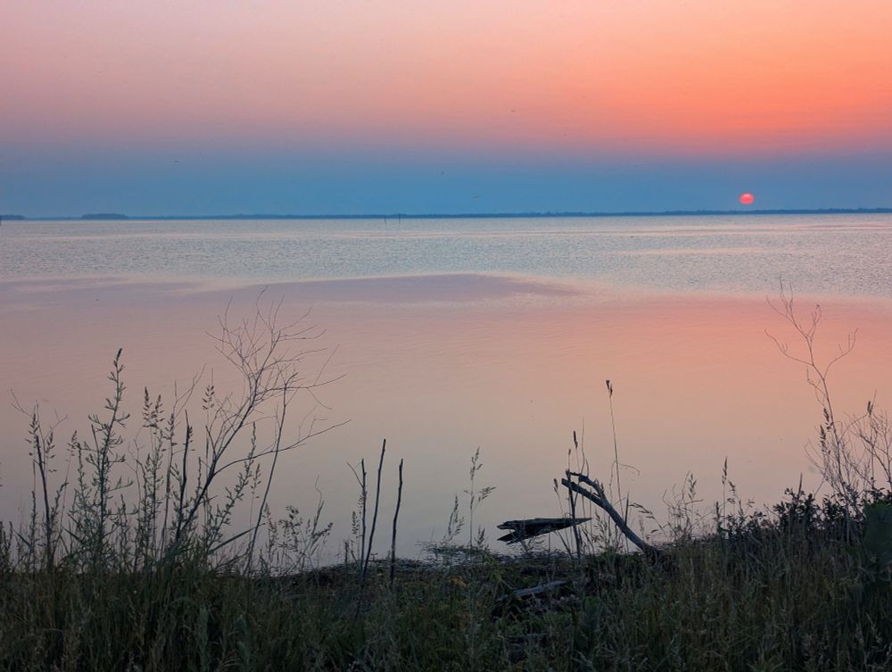 A fiery red dot of the sun rising over a flat calm lake with pastel oranges, red and pink reflect on the water 