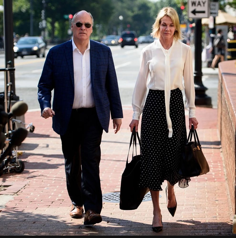 A picture of Craft and his wife walking down the street towards the camera. They are dressed in very expensive clothing. The wife is thin, w a blond bob. She's wearing a tasteful white blouse with an untied pussy bow, a mid length black skit w white dots, and the Carolyne shoe by Manolo Blahnik. She carries a Birkin bag. They both look angry and soulless.  