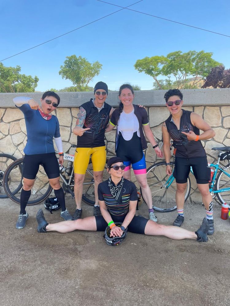 Group of cyclists posing with bikes, one cyclist is in the splits, bikes are behind them against a stone wall