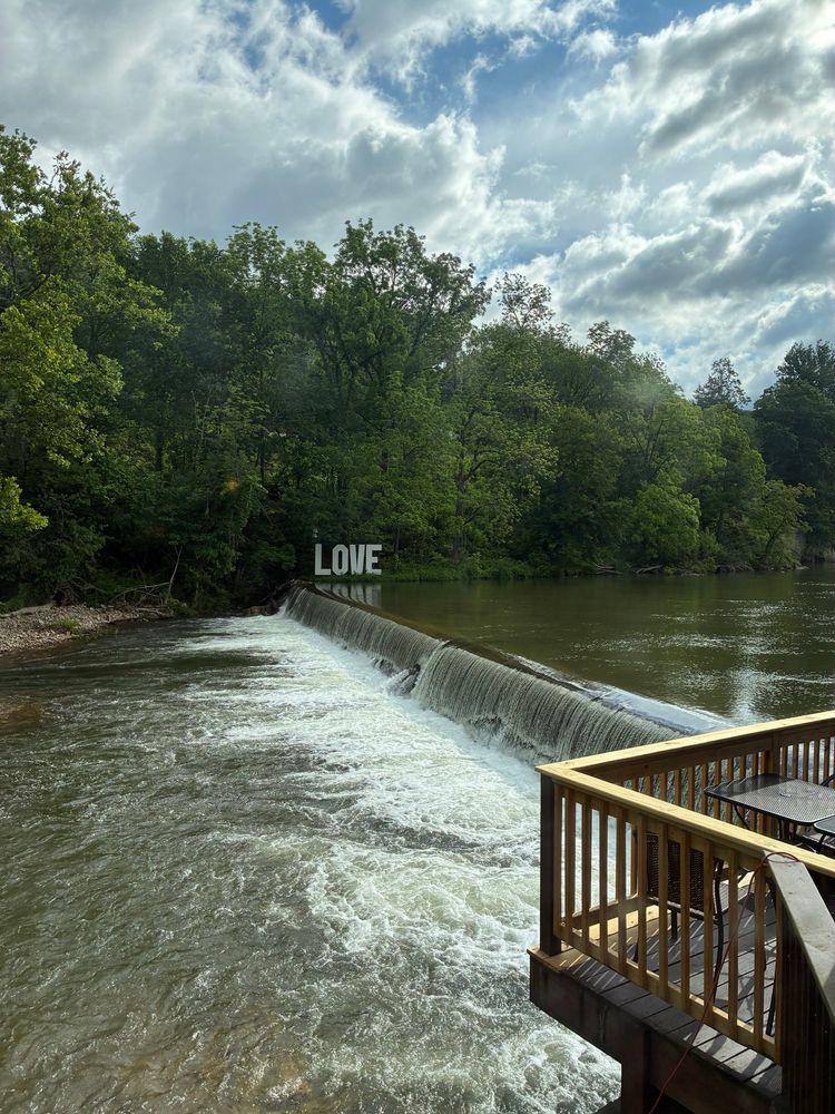 A peaceful river scene with a small waterfall flowing over a low dam surrounded by lush green trees under a partly cloudy sky. A wooden deck with patio furniture overlooks the water in the foreground. The word “LOVE” is prominently displayed on the opposite bank near the trees.