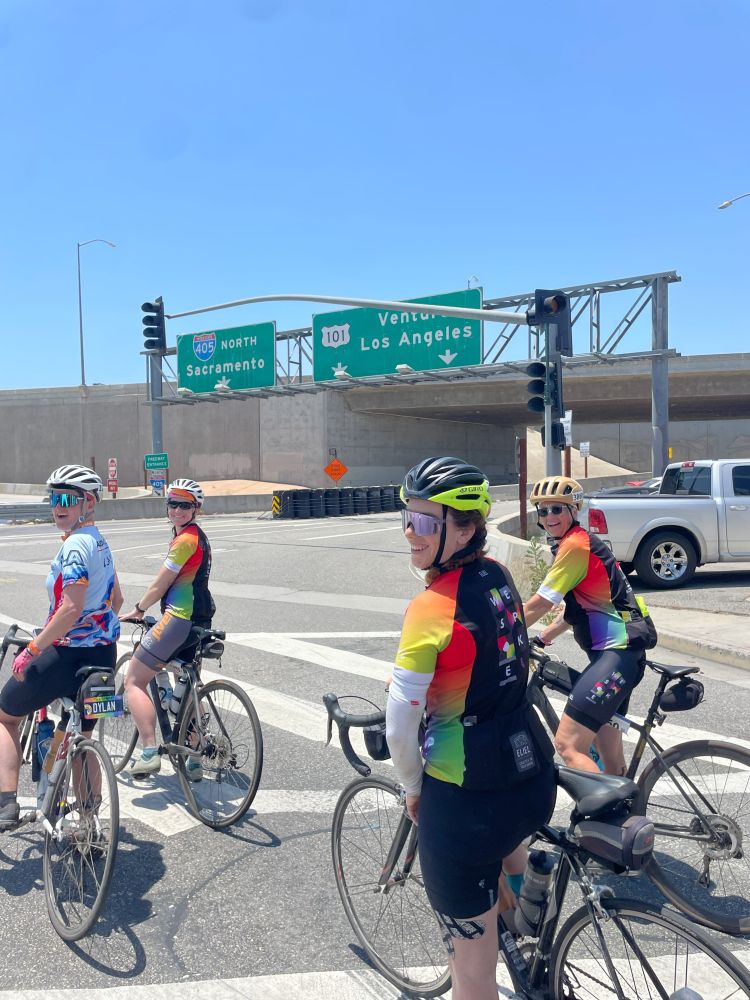 Group of cyclists in colorful jerseys on bikes crossing a highway, with highway signs for Sacramento and Los Angeles in the background