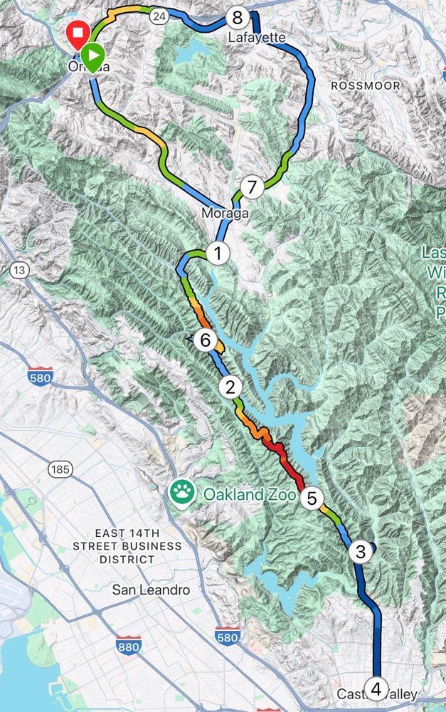 A digital map displaying a cycling route starting and ending in Orinda, California. The route passes through Moraga, Castro Valley, and Lafayette, covering varied terrain. Color-coded elevation markers indicate climbs and descents, with red and orange sections highlighting steep inclines. Nearby landmarks, including the Oakland Zoo, are visible.
