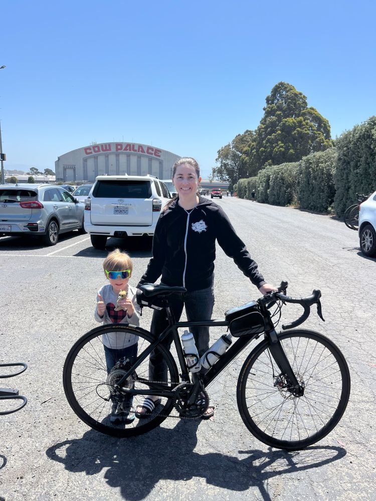 A person and a child stand proudly next to a black bicycle, with the Cow Palace venue in the background under a clear blue sky