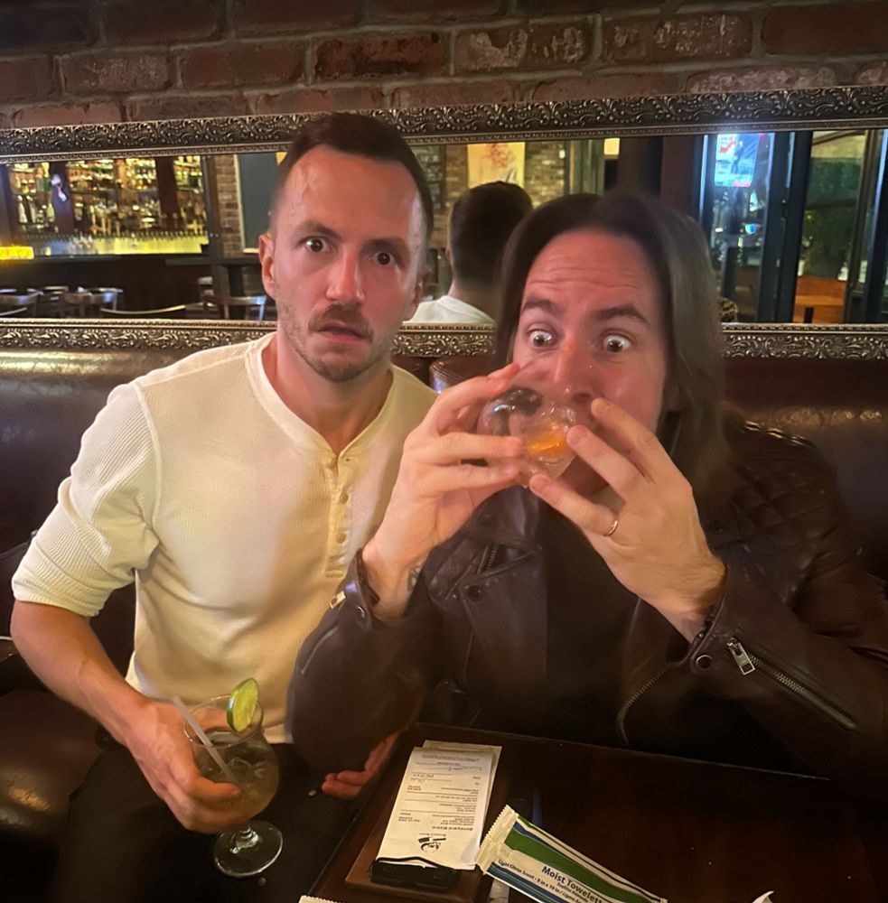 A photo of Alex Jordan and Matthew Mercer at a bar. Matthew is drinking a glass of alcohol with a gleeful expression and Alex is looking shocked/alarmed at the camera.
