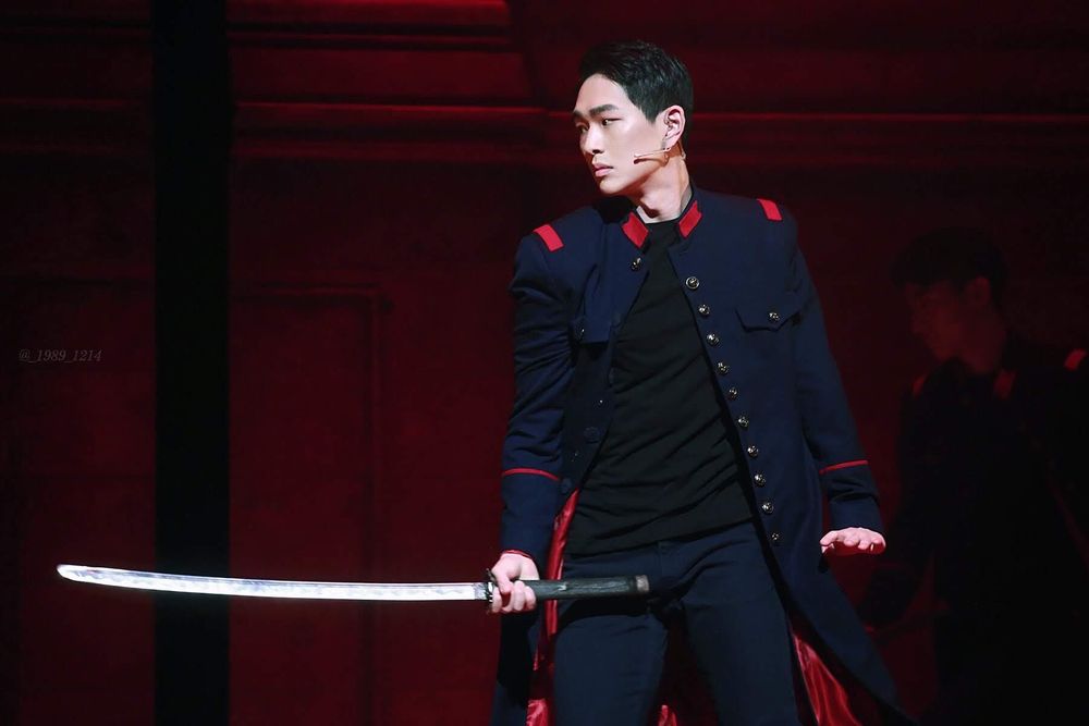 Jinki onstage during a 2019 performance of Shinheung Military Academy: The Musical. He’s wearing a dark blue military uniform with red accents and holding a long sword in his right hand as he looks to his right with a serious expression 