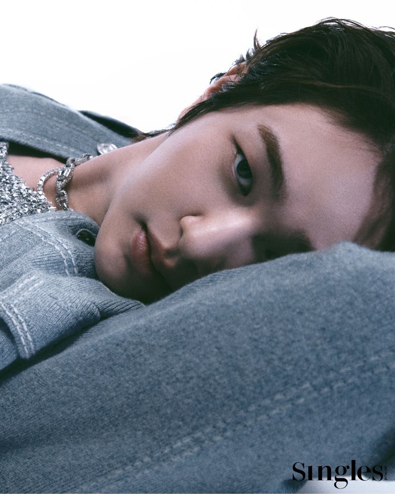 Jinki reclining with his arm behind his head, peeking at the camera over his elbow. He has dark hair and is wearing a blue-gray knit jacket and silver chains. 
Photo from a photoshoot for Singles magazine in 2024