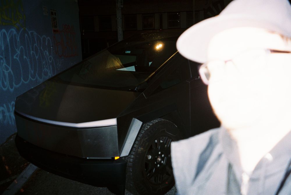 a flash light has overblown a person into whiteness, behind him is a cybertruck in the dark