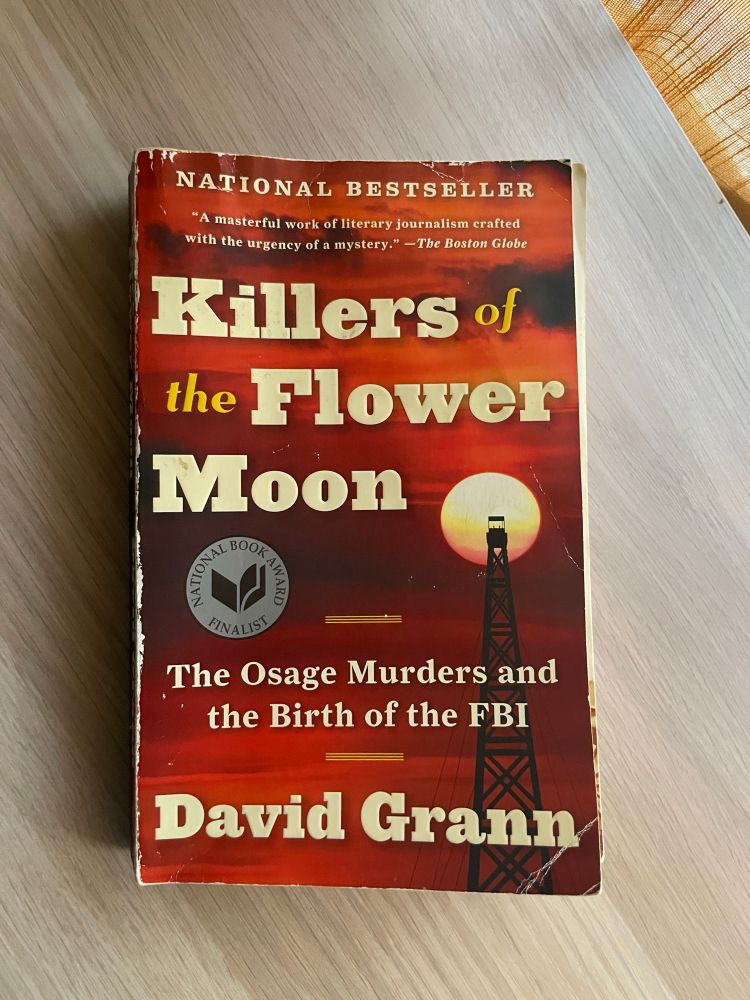 Killers of the Flower Moon by David Grann rests on a wooden surface. The cover is red and features an oil rig silhouetted against the sky. 