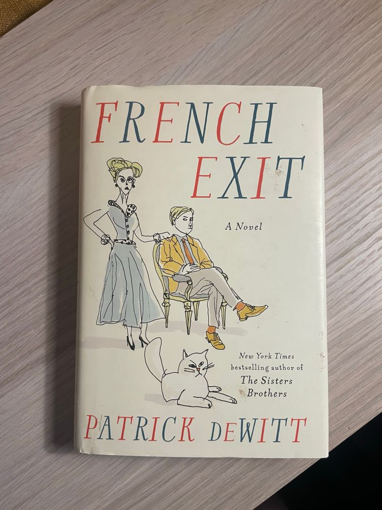 French Exit by Patrick deWitt rests on a wooden surface. The cover features an illustration of a man sitting in a chair with a woman standing behind him. The woman has her hand on his shoulder. A grumpy looking cat is on the floor in front of them.
