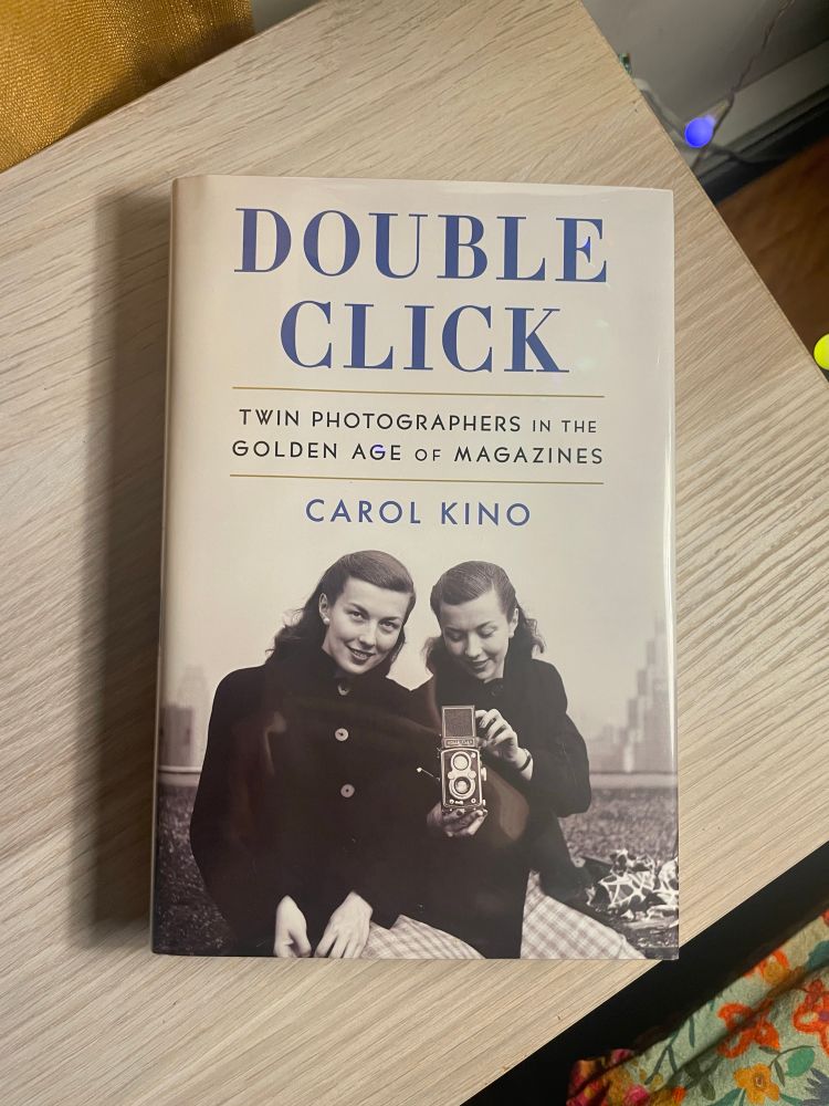 Double Click by Carol Kino rests on a wooden surface. The cover features a black-and-white photo of twin women. They have a shoulder length dark hair and are wearing dark coats. One of them is pointing a camera toward the unseen photographer.