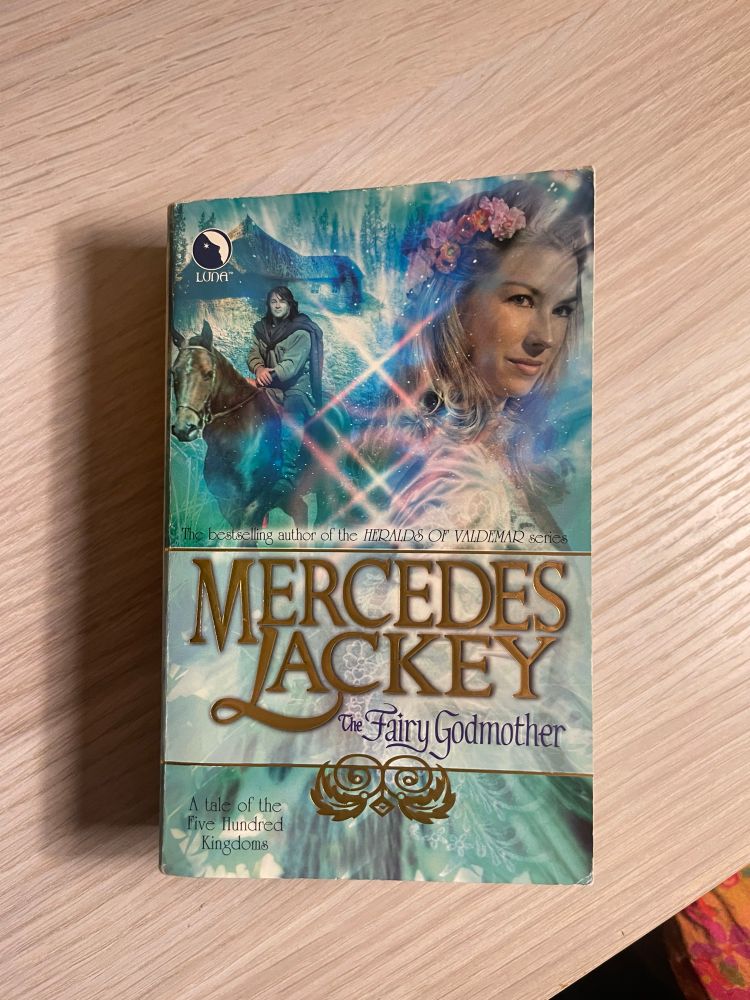 The Fairy Godmother by Mercedes Lackey rest on top of a wooden shelf. The cover features realistic illustrations of a woman in a beautiful gown with long blonde hair and a man with brown hair on a brown horse.