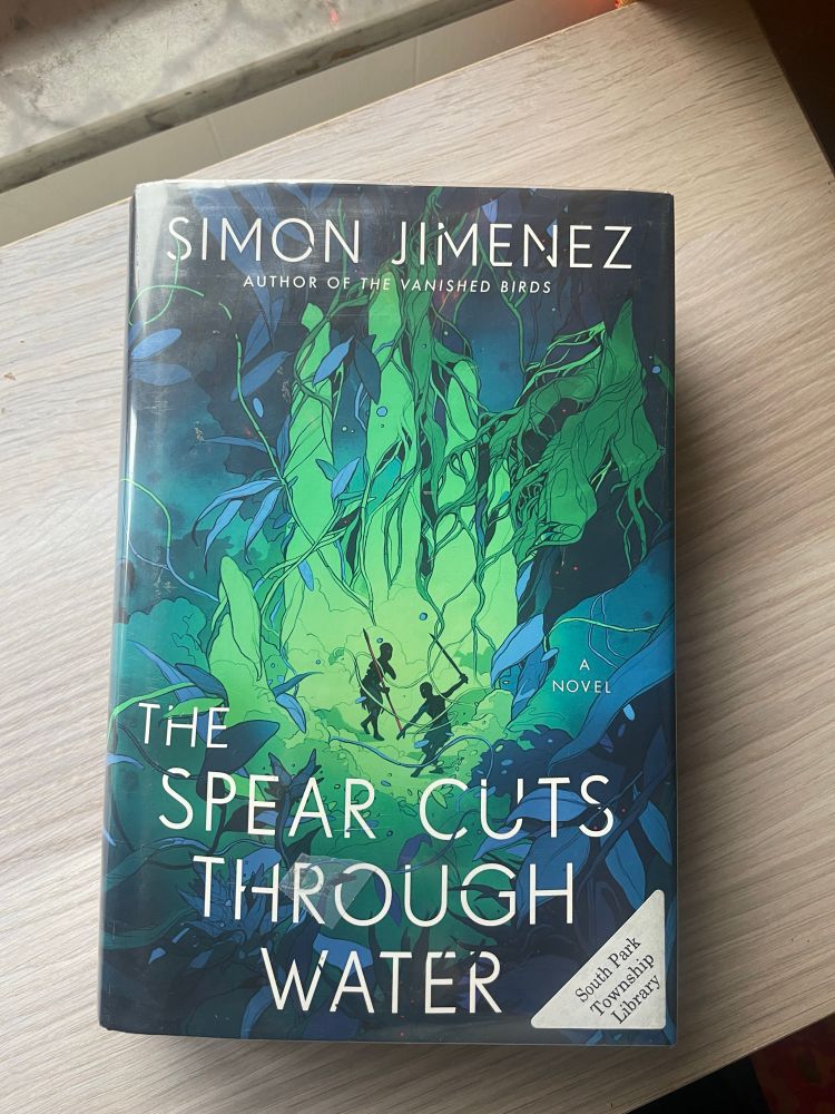 The Spear Cuts Through Water by Simon Jimenez rests on a wooden surface.