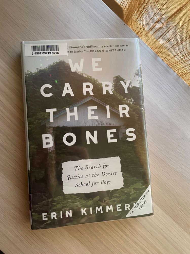 We Carry Their Bones by Erin Kimmerle rests on top of a wooden shelf. Cover features a photo of a dilapidated white building.