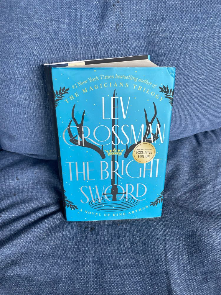 The Bright Sword by Lev Grossman sits on a blue couch. The cover is bright blue and features a sword, crown, and antlers. 