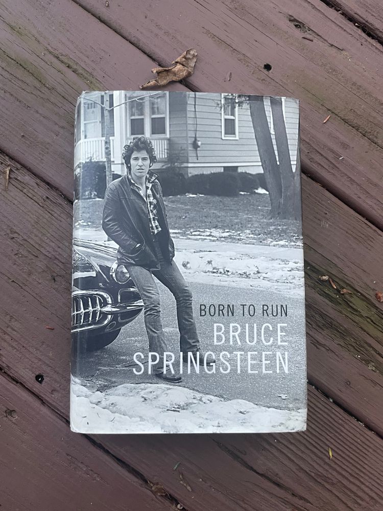 Born to Run by Bruce Springsteen rests on brown, wooden boards. Some dried leaves are scattered around the book. The cover features a black and white photo of The Boss as a young man leaning against the hood of a car. 
