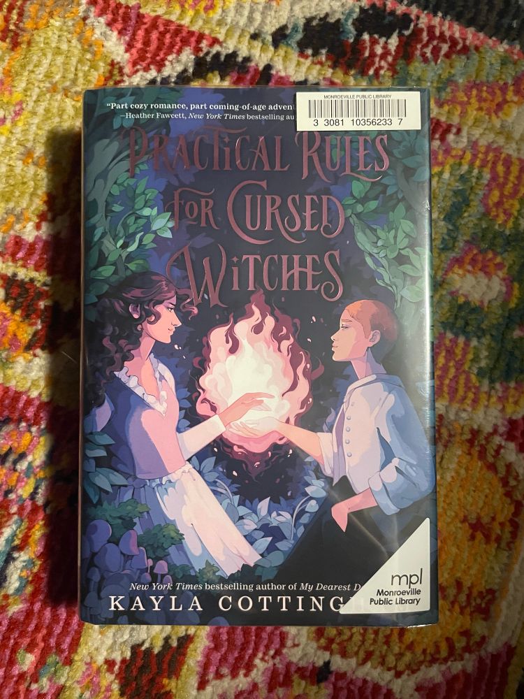 Practical Rules for Cursed Witches by Kayla Cottingham rests on a pink floral rug. The cover features an illustration of a tall witch with long curly brown hair and a shorter witch with short blonde hair facing each other. Fire burns between them, over their outstretched hands.