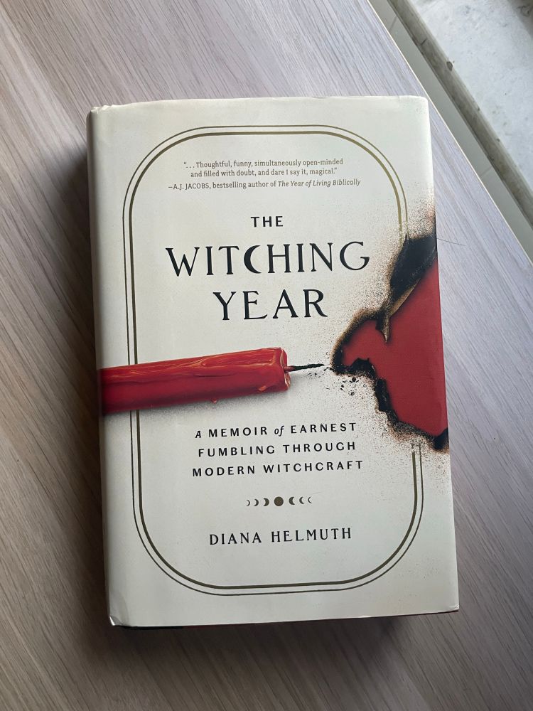 The Witching Year by Diana Helmuth rests on a wooden surface. The cover features and overturned red candle that is figuratively burning the cover of the book.