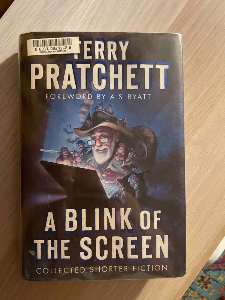 A Blink of the Screen by Terry Pratchett rests on top of a wooden shelf. The cover features an illustration of the author typing on a laptop surrounded by tiny characters from his stories.