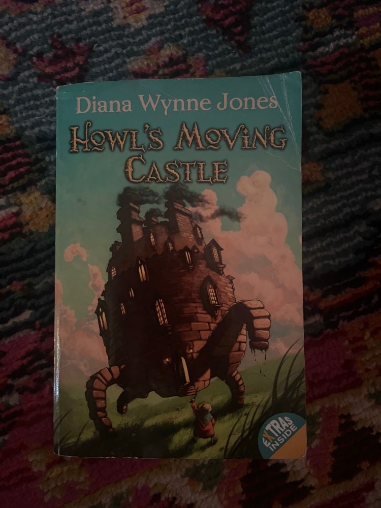 Howl’s Moving Castle by Diana Wynne Jones rests on a blue and pink floral rug. The cover features an illustration of a small round stone castle moving across a grassy landscape on four legs.