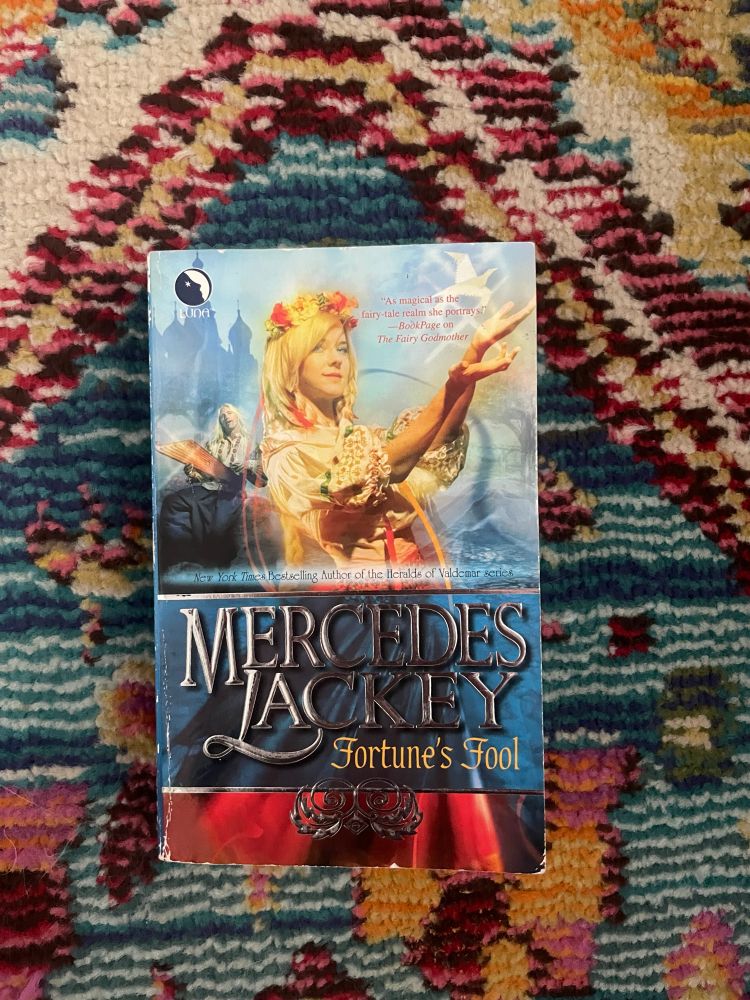 Fortune’s Fool by Mercedes Lackey rests on a pink and blue floral rug. The cover features an illustration of a young woman with long blonde hair and a flower crown, arms upraised.