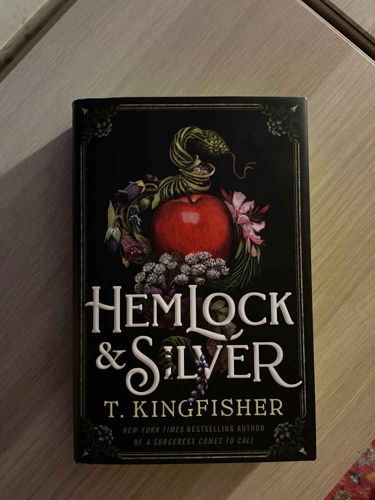 Hemlock & Silver by T. Kingfisher rests on top of a wooden shelf. The cover features a gorgeous illustration of an adder made of herbs and flowers coiling around a red apple. 