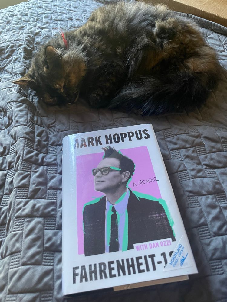 Fahrenheit-182 by Mark Hoppus and Dan Ozzi rests on a grey blanket. The cover features an image of Mark Hoppus, a white man in a suit. A fluffy calico cat is curled up above the book. 