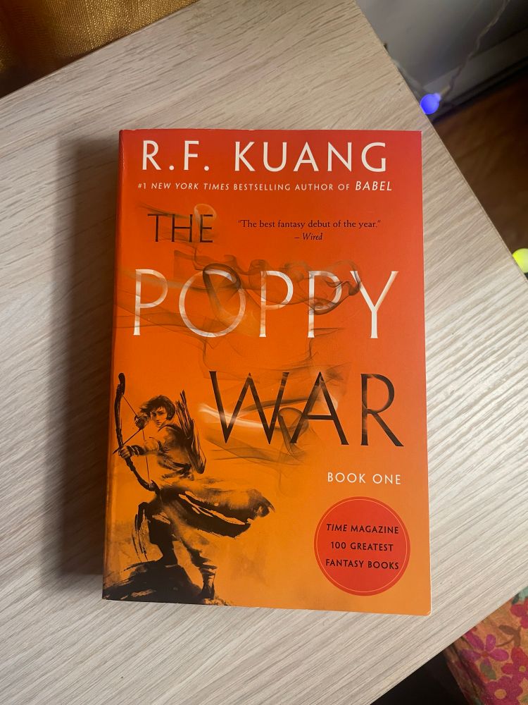The Poppy War by R.F. Kuang rests on a wooden surface. The book cover is orange and features a beautiful black-and-white illustration of a person in flowing robes with a bow and arrow.