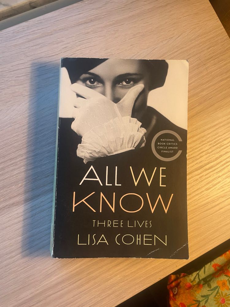 All We Know by Lisa Cohen rests on a wooden shelf. The cover features a black and white photo of a woman mischievously covering the lower half of her face with her hand. 