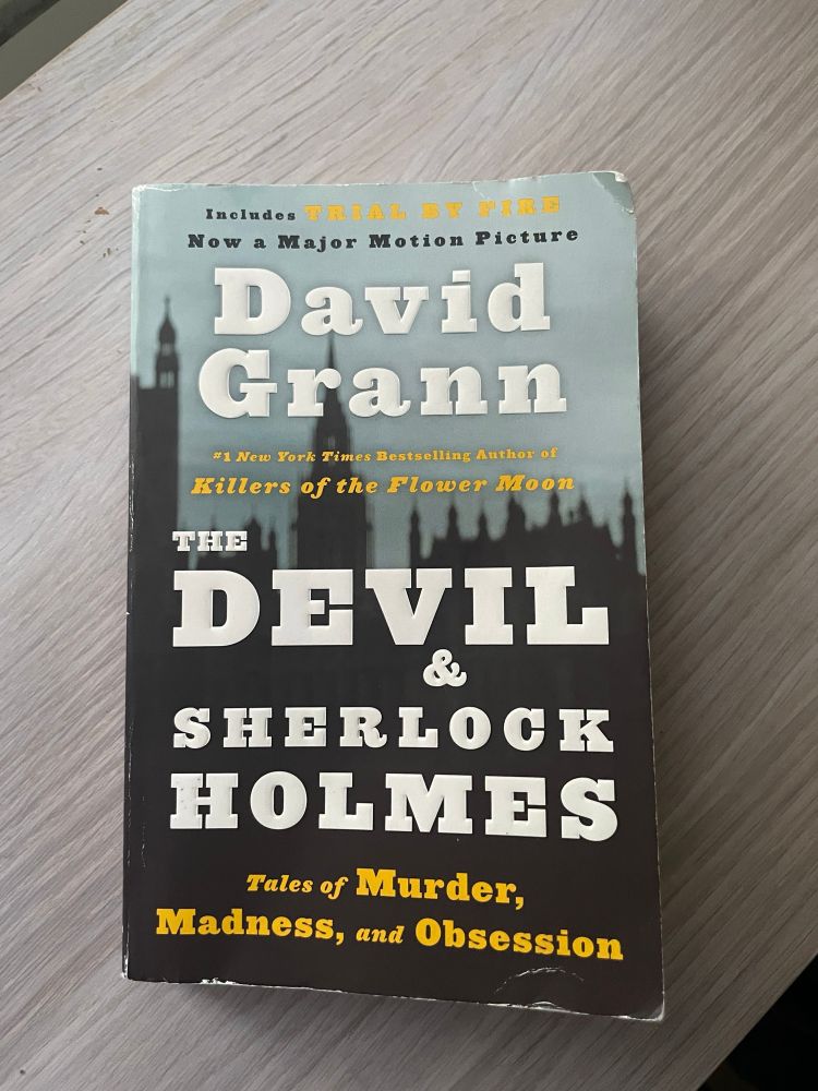 The Devil and Sherlock Holmes by David Grann rests on top of a wooden shelf.