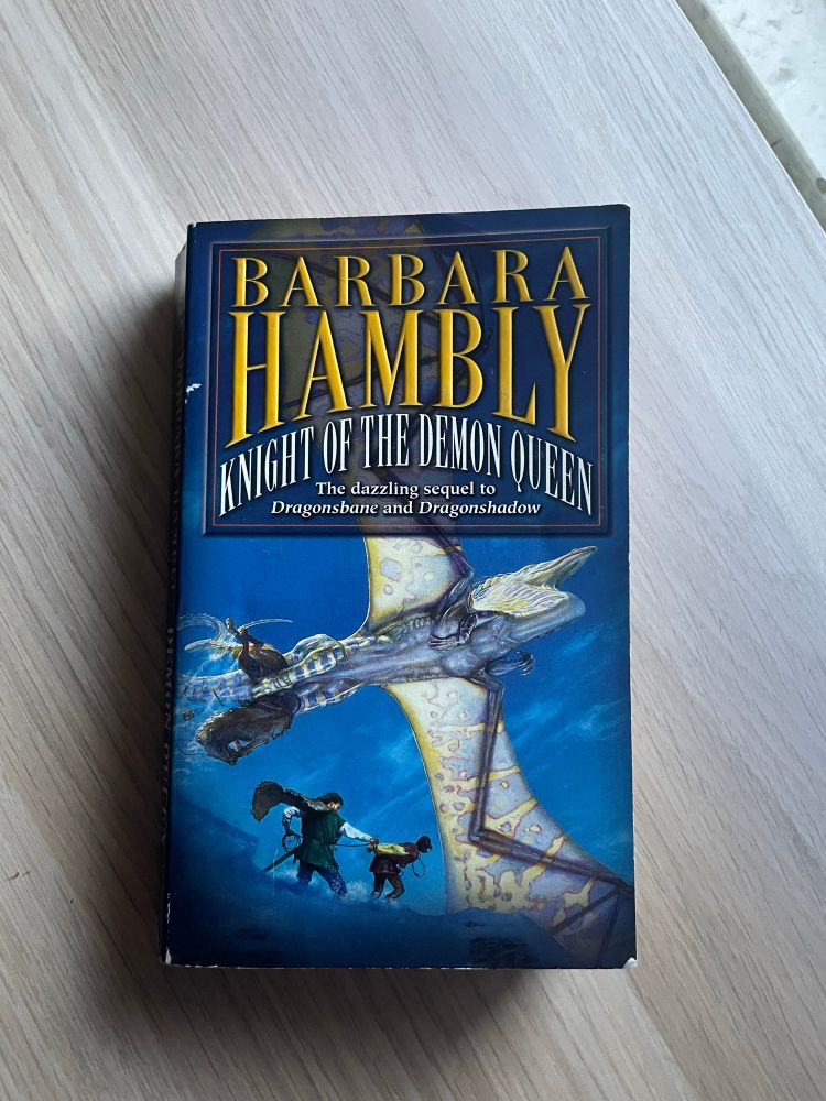 Knight of the Demon Queen by Barbara Hambly rests on a wooden surface. The cover features an illustration of a blue and gold dragon soaring above two small human figures.