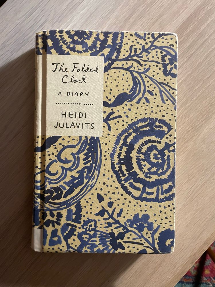 The Folded Clock: A Diary by Heidi Julavits rests on top of a faux wooden shelf. The cover has a beautiful design of blue flowers and plants. 
