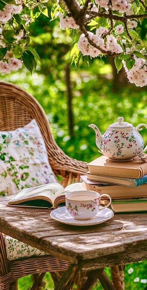 Sur cette photo nous voyons une table basse en bois brut une tasse de thé plusieurs livres et une théière en porcelaine à fleurs 
Un confortable fauteuil en osier garni d'un coussin à fleurs 
Le tout sur le gazon du jardin sous un cerisier en fleur 🌸 