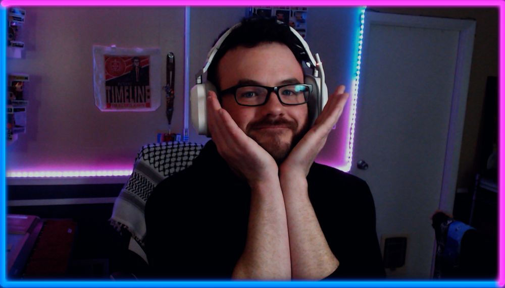 Jimmy with his two palms held under his chin with his fingers pointing upwards, wearing a white headset, a Keffiyeh on a chair in the background and blue and pink whites further in the background 