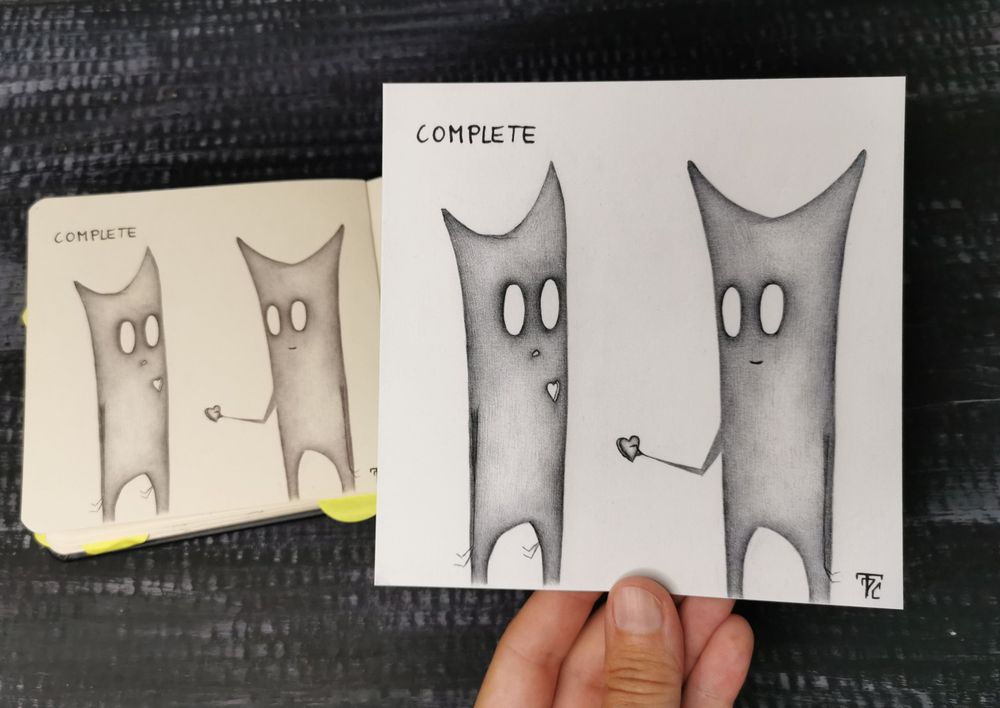 Auf dunklem Hintergrund liegt ein geöffnetes Skizzenbuch. Auf der linken Buchseite ist eine Zeichnung mit schwarzem Kugelschreiber. Das Motiv sind zwei geistartige Figuren mit großen, leeren Augen und zwei Spitzen am Kopf. Die linke Figur hat ein Loch in Herzform in der Brust. Die rechte Figur hält der anderen Figur lächelnd ein Herz hin. Links oben steht folgendes: Complete.
Eine Hand hält ein Kunstdruck über das Skizzenbuch. Das Motiv ist das gleiche.