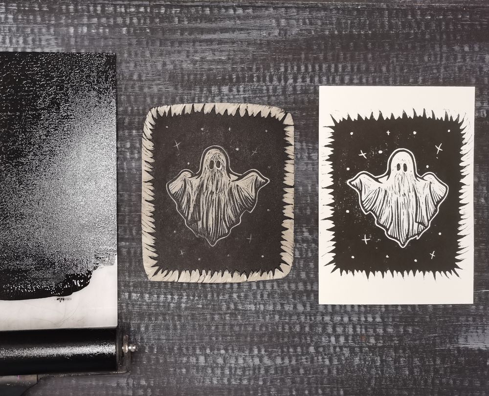 Auf dunklem Hintergrund liegt rechts ein Linoldruck mit schwarzer Farbe auf weißem Papier. Der Druck zeigt eine schwarze Fläche mit zackigem Rand. In der Mitte ist ein Geist zu sehen. Links daneben liegt der dazugehörige Linolblock mit schwarzer Farbe versehen. Weiter rechts daneben sieht man einen Teil einer Acrylplatte, auf der schwarze Farbe mittels einer Farbwalze ausgerollt wurde. 
